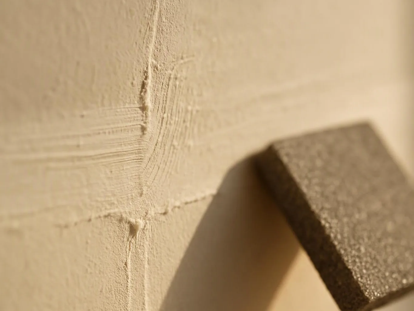 Close-up of a beige textured wall with a dark rectangular metal piece casting a soft shadow