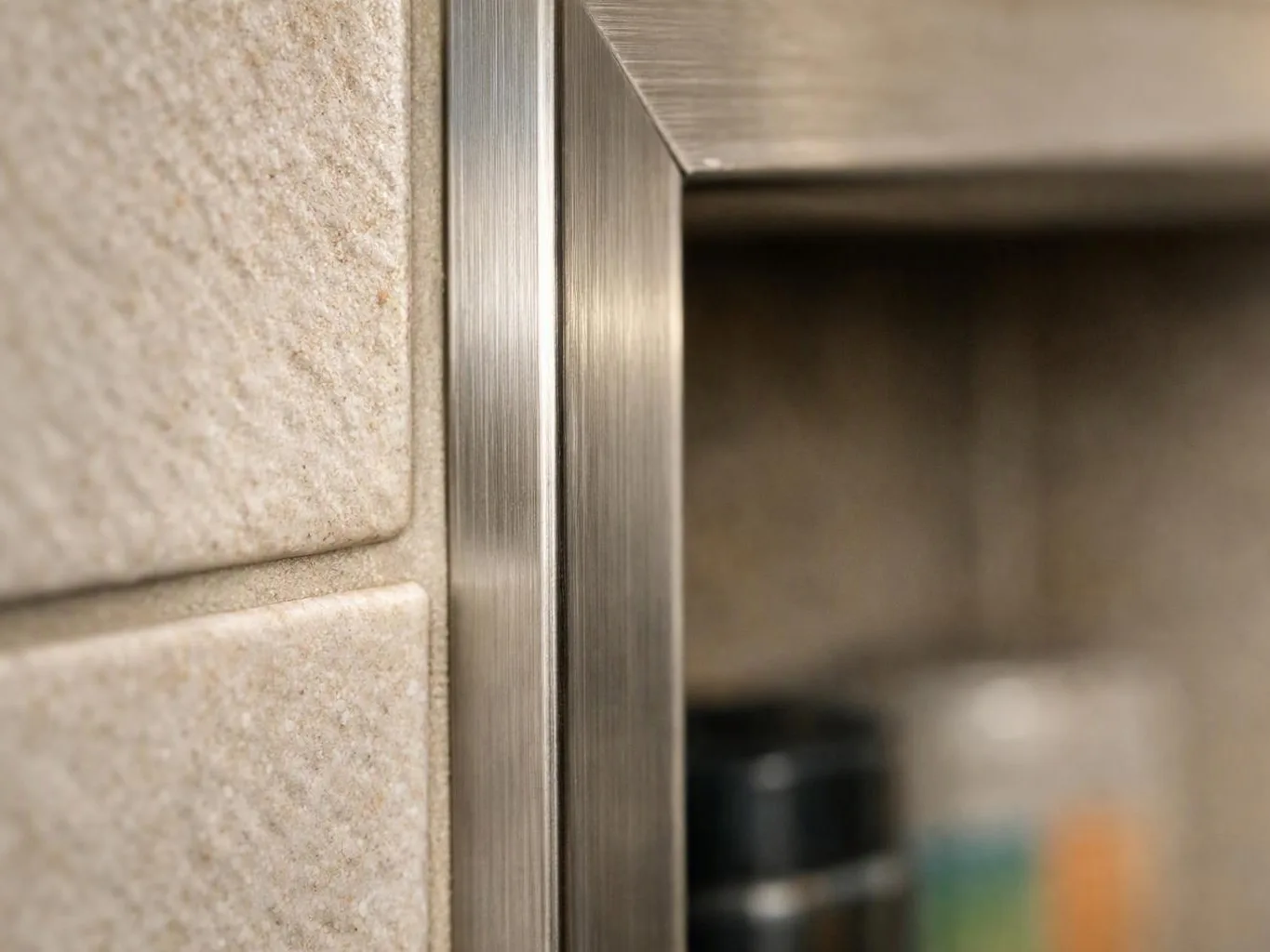 Close-up of a stainless steel cabinet frame beside textured beige tile, with a blurred shelf in the background