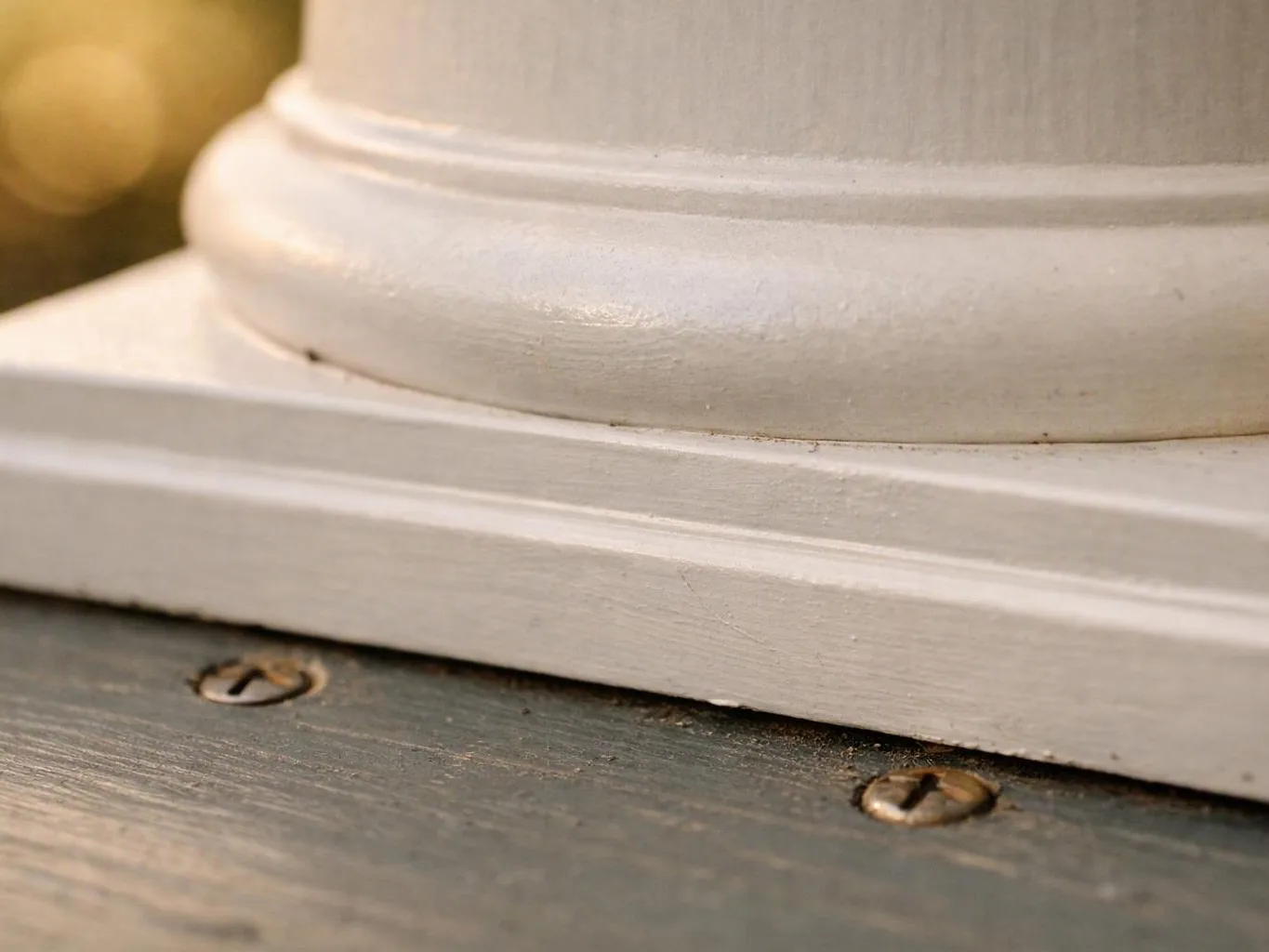 Close-up of a white painted column base on weathered wooden decking with visible screws and warm sunlight