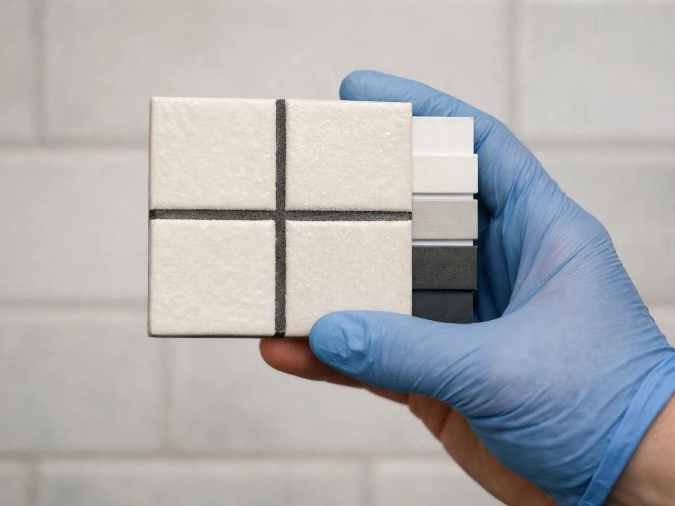 A gloved hand holds a square tile sample card with four light panels and stacked gray strips against a tiled wall