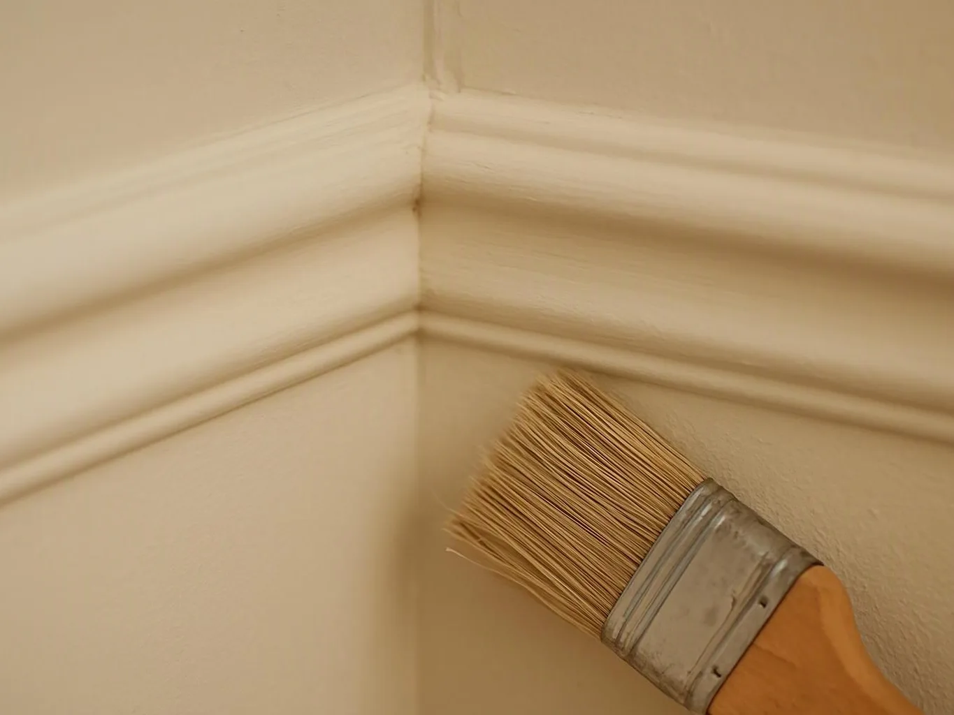 A paintbrush applies beige paint to a corner of a wall with white trim trim