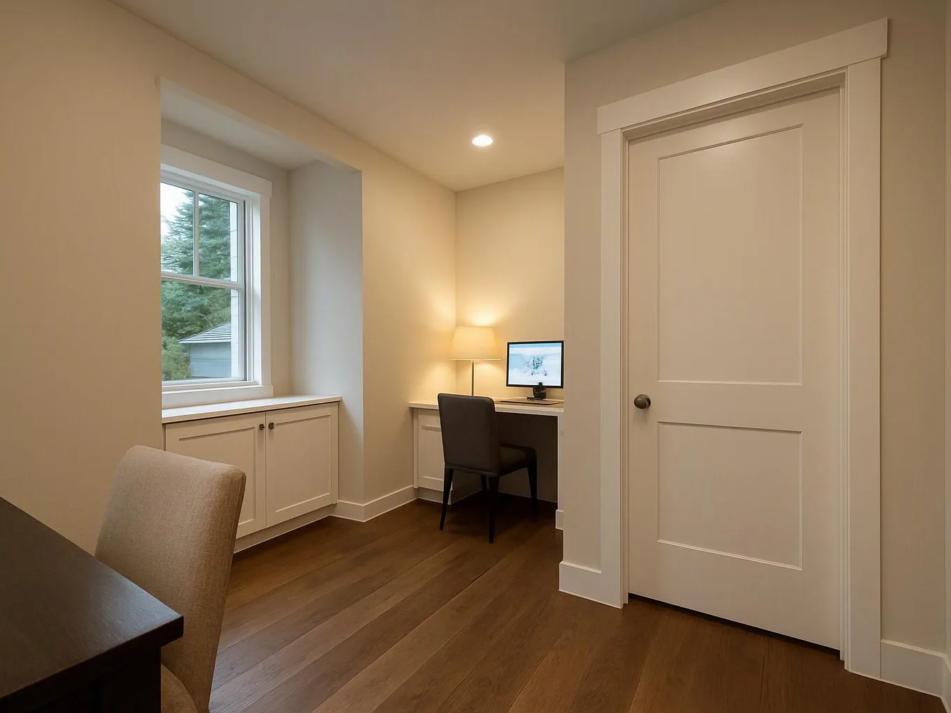 A small home office with a desk, chair, lamp, window, and closed door in warm light