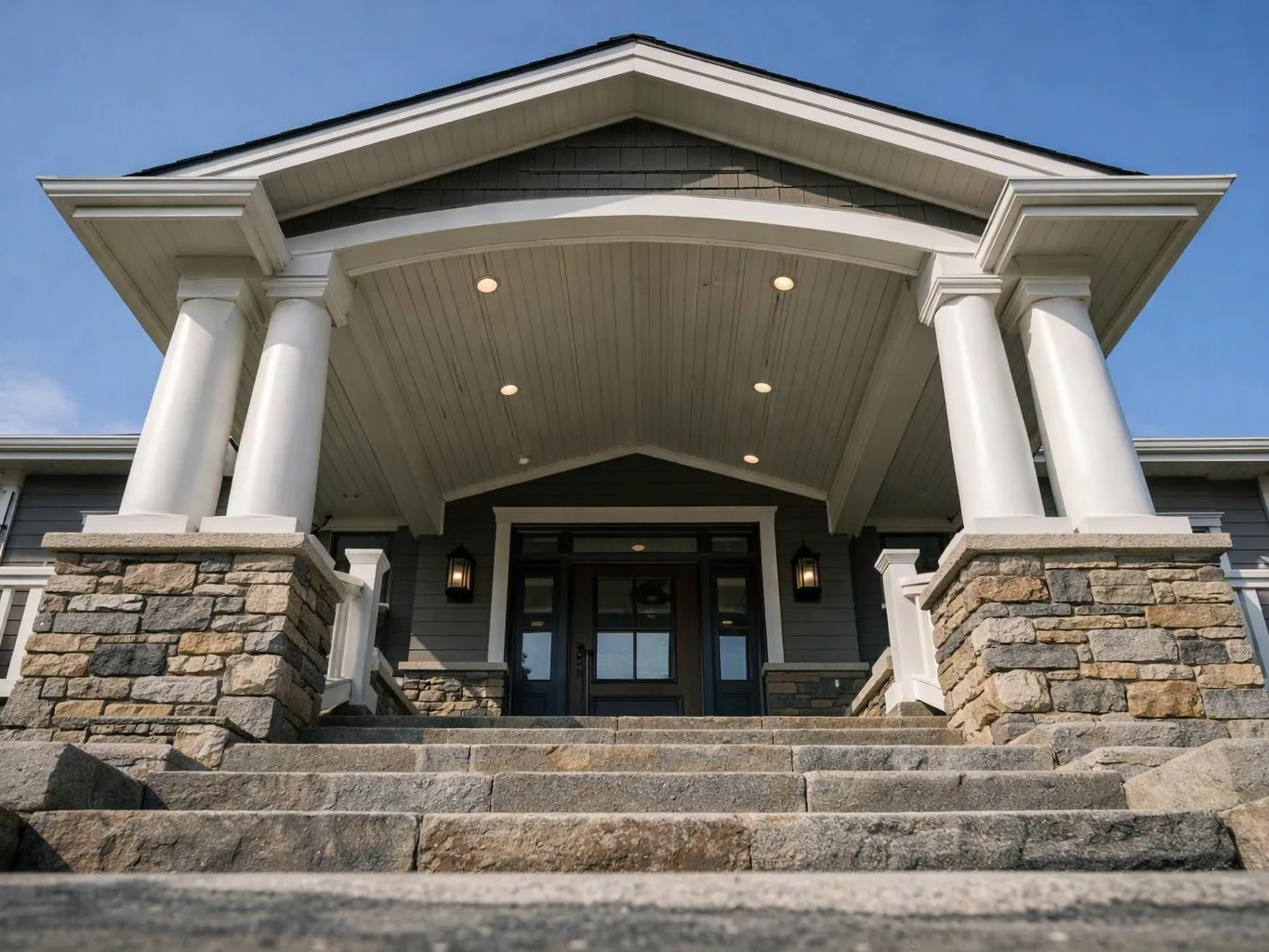 A stone staircase leads to a house entrance with white columns, a covered porch, and dark double doors