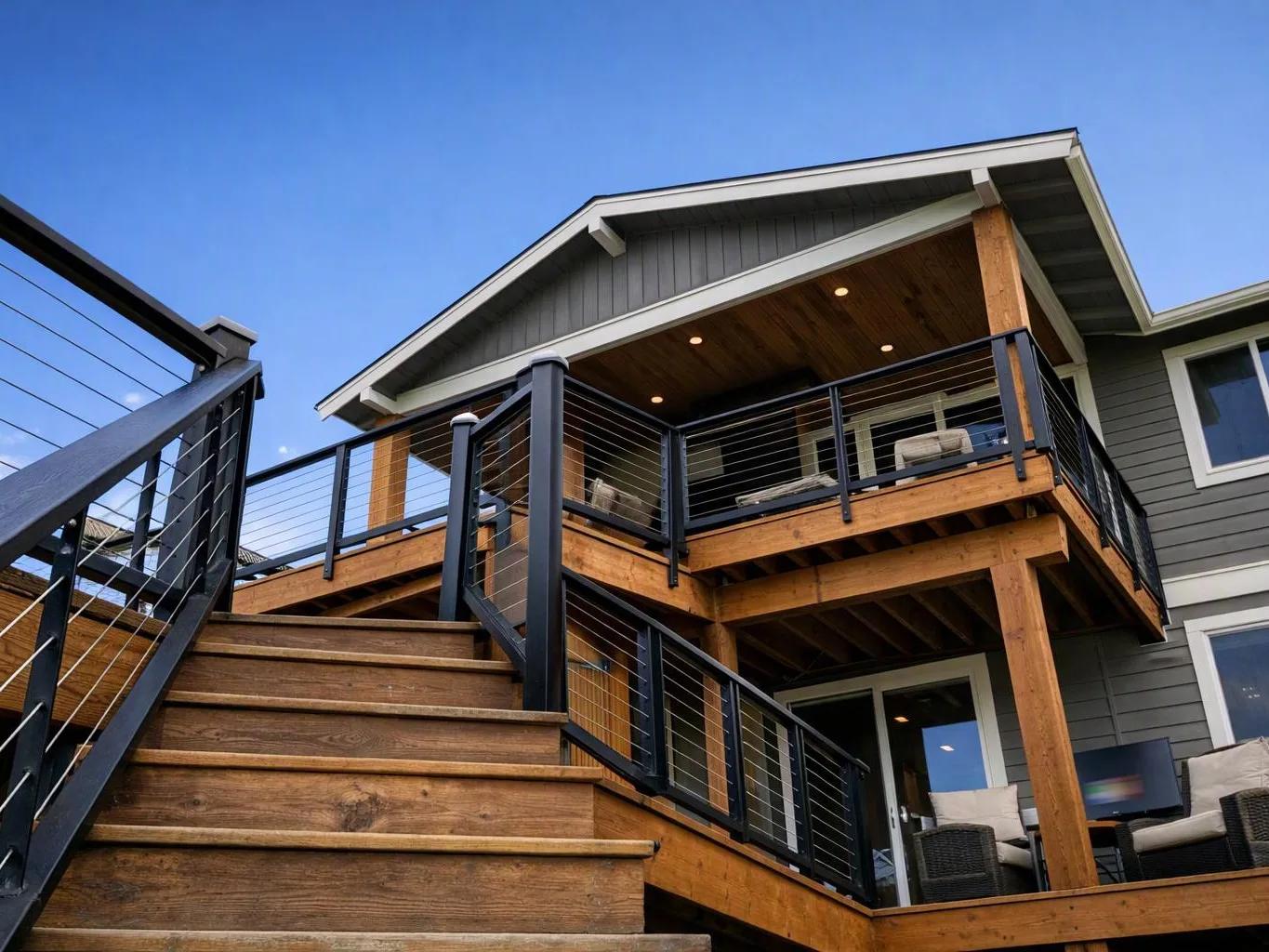 A wooden staircase leads up to a modern house deck with black railings under a clear blue sky